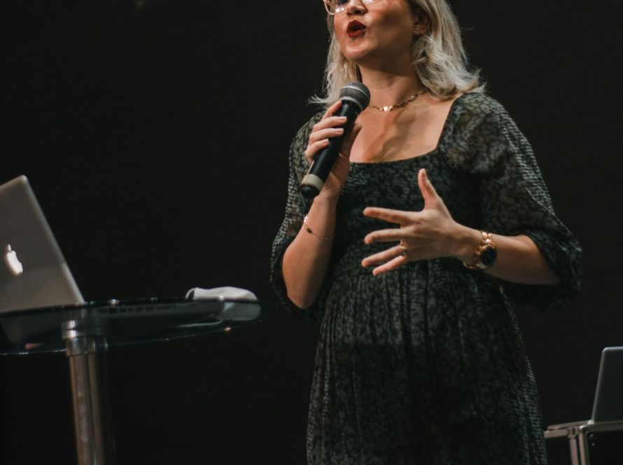 A person stands on stage speaking into a microphone at an event or presentation.